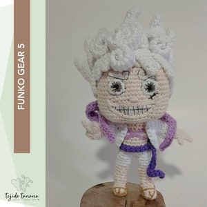 Könnte beinhalten: Eine gehäkelte Funko Pop Figur eines Charakters mit weißen Haaren, einem lila und weißen Outfit und einem verschmitzten Grinsen. Die Figur steht auf einem Holzsockel. Der Text "FUNKO GEAR 5" ist auf der linken Seite des Bildes sichtbar.