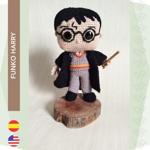 Puede incluir: Figura de Harry Potter Funko Pop hecha a crochet con una túnica negra, pantalones grises y una bufanda roja y amarilla. La figura sostiene una varita mágica y está de pie sobre una base de madera.