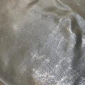 May include: A close-up of a smooth, silver-coloured fabric with a subtle sheen. The fabric has a slightly wrinkled texture, creating a soft, flowing appearance.