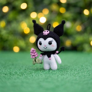 Pode incluir: Um brinquedo de pelúcia branco de crochê com orelhas pretas e uma caveira preta na cabeça. O brinquedo está segurando uma flor rosa e está em pé na grama verde.