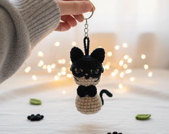 May include: A handmade crochet cat keychain with a black head, a beige body, and a black bow tie. The cat has round eyes and is held by a silver keyring. Black paw-shaped decorations and green leaf-shaped decorations are scattered around the cat.