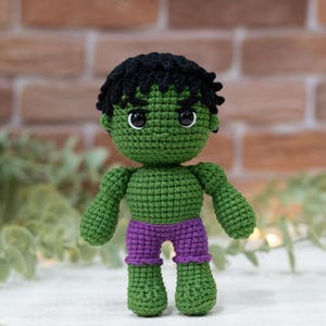 Puede incluir: Muñeco de Hulk hecho a mano con ganchillo. El muñeco es principalmente verde, con pelo negro y pantalones cortos morados. El muñeco tiene un aspecto detallado y texturizado, con puntadas visibles. Los ojos son negros y blancos. El fondo es una pared de ladrillos.