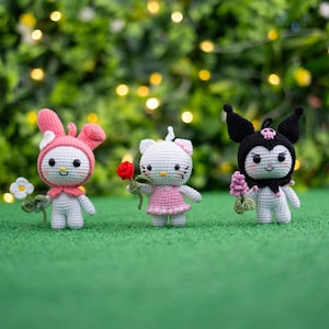 May include: Three crocheted plush toys, a pink bunny holding a white flower, a white cat holding a red rose, and a black and white cat with bat ears holding a purple flower. The toys are sitting on a green surface.