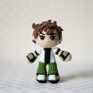Puede incluir: Un juguete de peluche tejido a crochet de un personaje de dibujos animados con cabello castaño, ojos verdes y una camisa a rayas blancas y negras. El juguete lleva pantalones verdes y brazaletes verdes y negros.