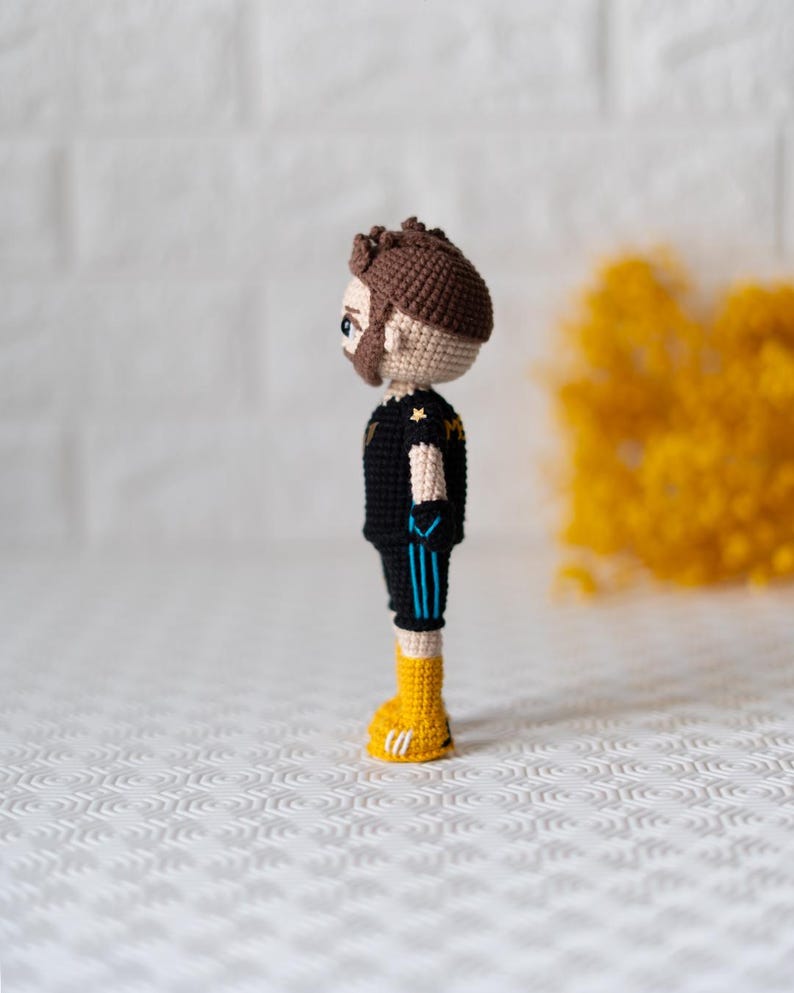 May include: A crocheted figurine of a person wearing a black and blue jersey with a yellow star on the chest, black shorts with blue stripes, and yellow shoes.