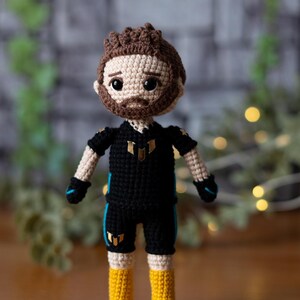 May include: Crocheted doll with brown hair, a black shirt with gold accents, and yellow boots. The doll is wearing a black and blue outfit with a gold logo.