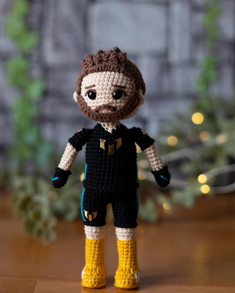 May include: Crocheted doll with brown hair, a black shirt with gold accents, and yellow boots. The doll is wearing a black and blue outfit with a gold logo.