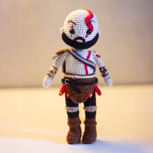 Pode incluir: Boneca de croch&ecirc; feita &agrave; m&atilde;o de um personagem com cabe&ccedil;a branca, barba preta e marcas vermelhas. A boneca usa uma roupa marrom e preta com detalhes vermelhos, botas marrons e uma al&ccedil;a no peito. A boneca est&aacute; em p&eacute;.