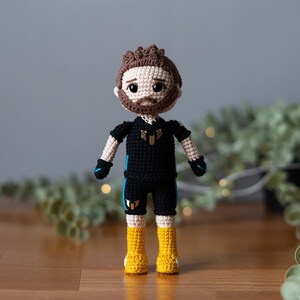 May include: Crocheted doll with brown hair, a black shirt with gold accents, and yellow boots. The doll is standing on a wooden surface.