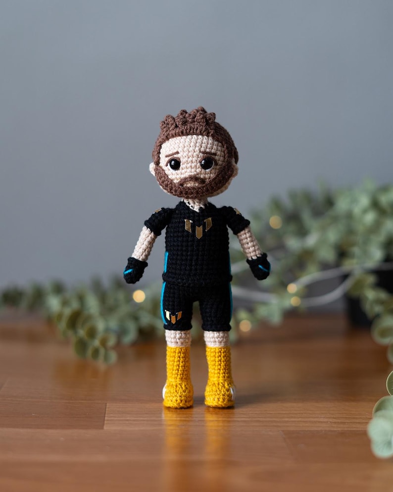 May include: Crocheted doll with brown hair, a black shirt with gold accents, and yellow boots. The doll is standing on a wooden surface.