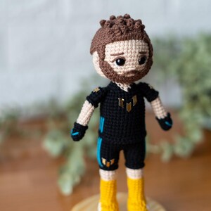 May include: A crocheted doll wearing a black and blue jersey with gold accents. The doll has brown hair, a beard, and black eyes. The doll is standing on a wooden base.