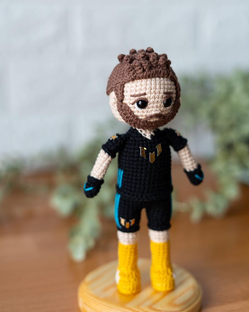 May include: A crocheted doll wearing a black and blue jersey with gold accents. The doll has brown hair, a beard, and black eyes. The doll is standing on a wooden base.