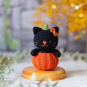 May include: A handmade crochet black cat wearing a green stem and orange bow, sitting inside an orange pumpkin. The cat has a smiling face with whiskers. The Halloween-themed cat is displayed on a small wooden stand.
