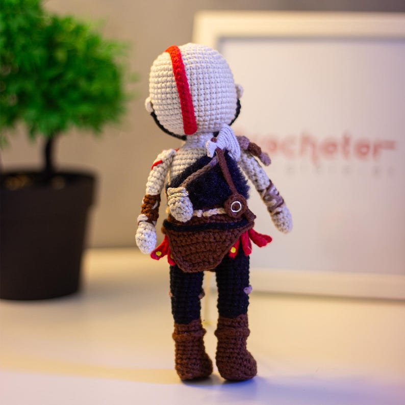 Pode incluir: Boneca de croch&ecirc; feita &agrave; m&atilde;o de um personagem com cabe&ccedil;a branca, uma faixa vermelha e detalhes pretos. A boneca usa uma bolsa marrom, cal&ccedil;as pretas e botas marrons. Detalhes vermelhos s&atilde;o vis&iacute;veis nas roupas da boneca.