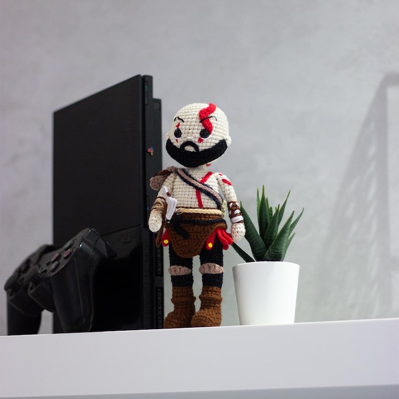 Pode incluir: Boneca de croch&ecirc; feita &agrave; m&atilde;o de um personagem de videogame, com cabe&ccedil;a branca, listra vermelha, barba preta e roupas marrons e pretas. A boneca est&aacute; em uma prateleira branca ao lado de uma pequena planta em vaso e um console de jogos preto.