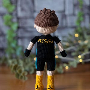 May include: A crocheted doll wearing a black and blue outfit with the text "M:55" on the front. The doll has brown hair and is wearing yellow boots.