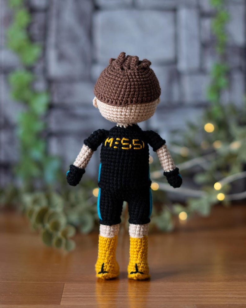 May include: A crocheted doll wearing a black and blue outfit with the text "M:55" on the front. The doll has brown hair and is wearing yellow boots.