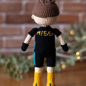 May include: Crocheted doll wearing a black and blue jersey with the word "MESSI" on the front. The doll has brown hair and is wearing yellow boots.