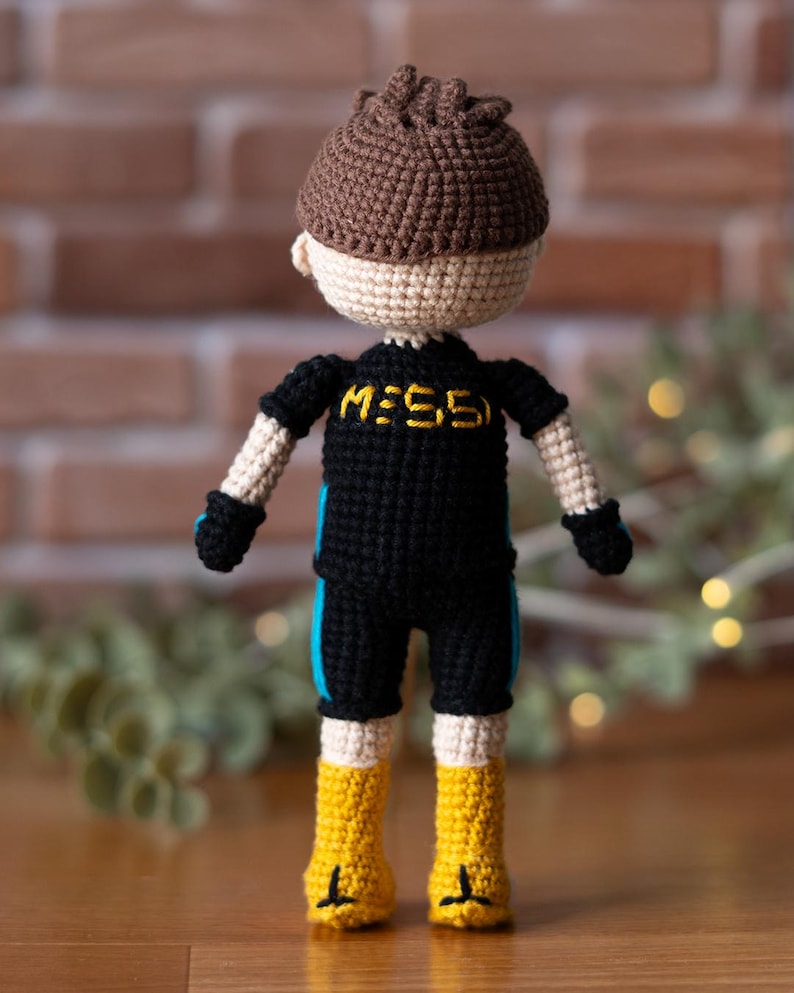 May include: Crocheted doll wearing a black and blue jersey with the word "MESSI" on the front. The doll has brown hair and is wearing yellow boots.