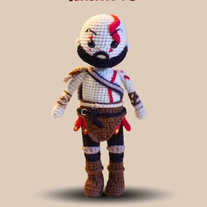 Pode incluir: Boneca amigurumi de croch&ecirc; de Kratos, personagem do videogame God of War. A boneca tem uma cabe&ccedil;a branca com uma faixa vermelha, barba preta e est&aacute; vestindo roupas marrons e pretas. O texto "AMIGURUMI PATTERN KARATOS" est&aacute; no topo.