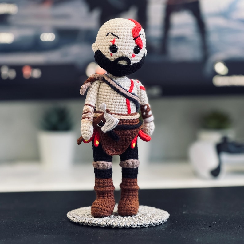 Pode incluir: Figura de croch&ecirc; de um personagem com cabe&ccedil;a branca, barba preta e marcas vermelhas. A figura usa uma roupa bege e marrom com um machado marrom e uma base circular. A figura tem aproximadamente 15 cm de altura.
