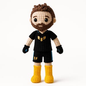 Puede incluir: Un juguete de peluche tejido a crochet que representa a una persona con un jersey negro con un logotipo dorado, pantalones cortos negros con ribetes azules y botas amarillas. El juguete tiene el pelo castaño y barba.