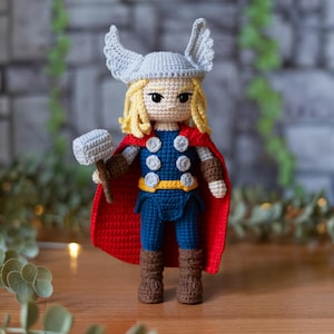 Könnte beinhalten: Handgemachte Häkelpuppe von Thor, mit silbernem Helm mit Flügeln, blondem Haar und rotem Umhang. Die Puppe trägt ein blau-weißes Outfit mit braunen Stiefeln und hält einen grauen Hammer. Die Puppe ist etwa 20 cm groß.
