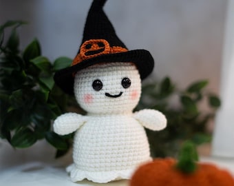 May include: A white crocheted ghost wearing a black witch hat with an orange pumpkin design. The ghost has black eyes and a smiling mouth. The ghost is sitting next to a small orange pumpkin.
