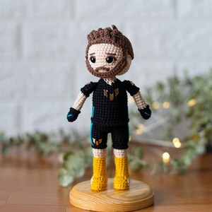 May include: Crocheted doll with brown hair, a black and yellow outfit, and yellow boots. The doll is standing on a wooden base.