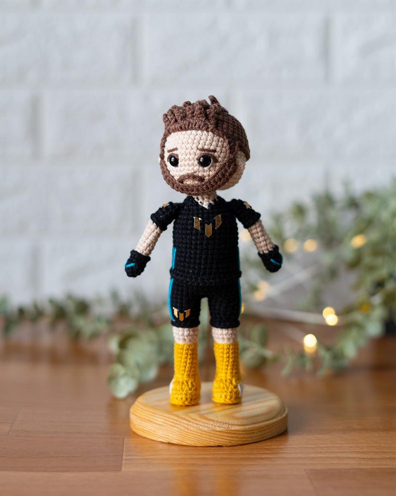 May include: Crocheted doll with brown hair, a black and yellow outfit, and yellow boots. The doll is standing on a wooden base.