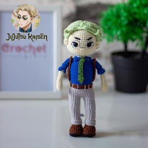 May include: A crocheted doll with green hair, blue shirt, green tie, brown trousers, and brown shoes. The doll is wearing glasses and has a serious expression. The doll is standing on a white surface. The image also includes the text "Jujutsu Kaisen Crochet".
