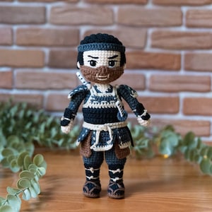 Könnte beinhalten: Handgefertigte Häkelpuppe eines Samurai-Kriegers, mit einem schwarz-blauen Outfit mit braunen Akzenten. Die Puppe hat ein detailliertes Gesicht mit einem braunen Bart und einem Stirnband. Die Puppe steht auf einer Holzoberfläche.