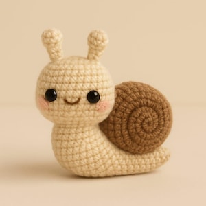 Könnte beinhalten: Eine gehäkelte Schneckenfigur mit cremefarbenem Körper und braunem Gehäuse. Die Schnecke hat schwarze Augen, rosige Wangen und kleine Fühler. Das handgefertigte Plüschtier ist ein niedliches und skurriles Dekorationsstück.