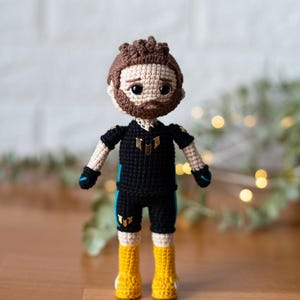May include: Crocheted amigurumi doll with brown hair, a black shirt with blue accents, and yellow boots. The doll is wearing a black and white outfit with a gold symbol on the chest.