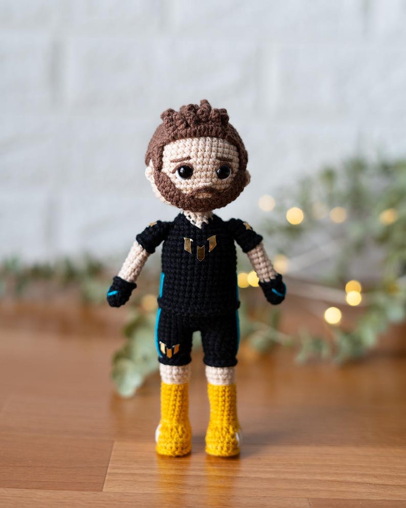 May include: Crocheted amigurumi doll with brown hair, a black shirt with blue accents, and yellow boots. The doll is wearing a black and white outfit with a gold symbol on the chest.