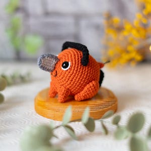 以下が含まれることがあります： 手作りのオレンジ色の編みぐるみで、灰色の鼻と黒いディテールがあります。小さな木製の台座の上に置かれています。頭には黒いチェーンソーのようなディテールがあり、小さな黒い尾が付いています。