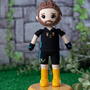 May include: Crocheted doll with brown hair, a beard, and a black and blue jersey with a gold emblem. The doll is wearing yellow boots and is standing on a wooden base.