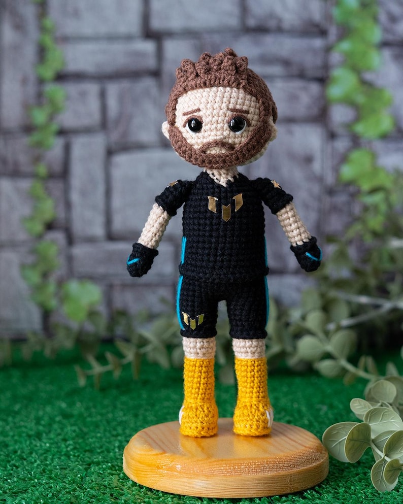 May include: Crocheted doll with brown hair, a beard, and a black and blue jersey with a gold emblem. The doll is wearing yellow boots and is standing on a wooden base.
