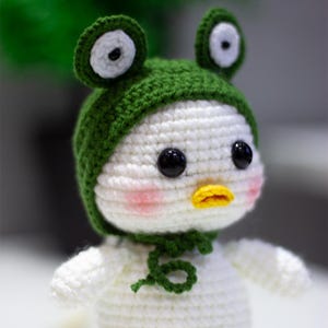May include: A white crocheted stuffed animal with a green frog hat. The animal has black eyes, a yellow beak, and pink cheeks.