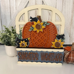 May include: A decorative wooden sign featuring an arched window frame, a central orange pumpkin with a lattice pattern, three black birds, and three sunflowers with button centers. The base is blue with white polka dots and the text SWEATER WEATHER.