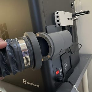 Könnte beinhalten: Ein schwarzer flexibler Schlauch ist mit einem grauen zylindrischen Adapter verbunden, der mit einer Metallklemme befestigt ist. Der Adapter ist an einer schwarzen Maschine mit dem Logo "Bambu Lab" befestigt. Ein weißes Gerät mit vier kreisförmigen Öffnungen ist an der Maschine montiert.