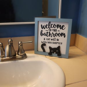 May include: A framed sign with a light blue border that reads "welcome to the bathroom" with a black cat graphic. The sign is on a bathroom counter near a sink and faucet.