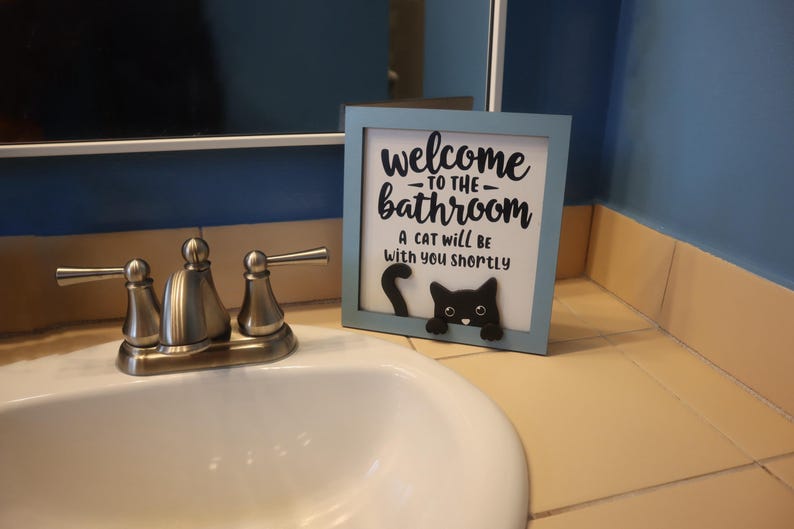May include: A framed sign with a light blue border that reads "welcome to the bathroom" with a black cat graphic. The sign is on a bathroom counter near a sink and faucet.