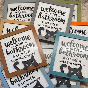 May include: A collection of framed bathroom signs with a playful cat design. Each sign features the text "Welcome to the Bathroom" and "A cat will be with you shortly" in black lettering. The frames come in various colors.