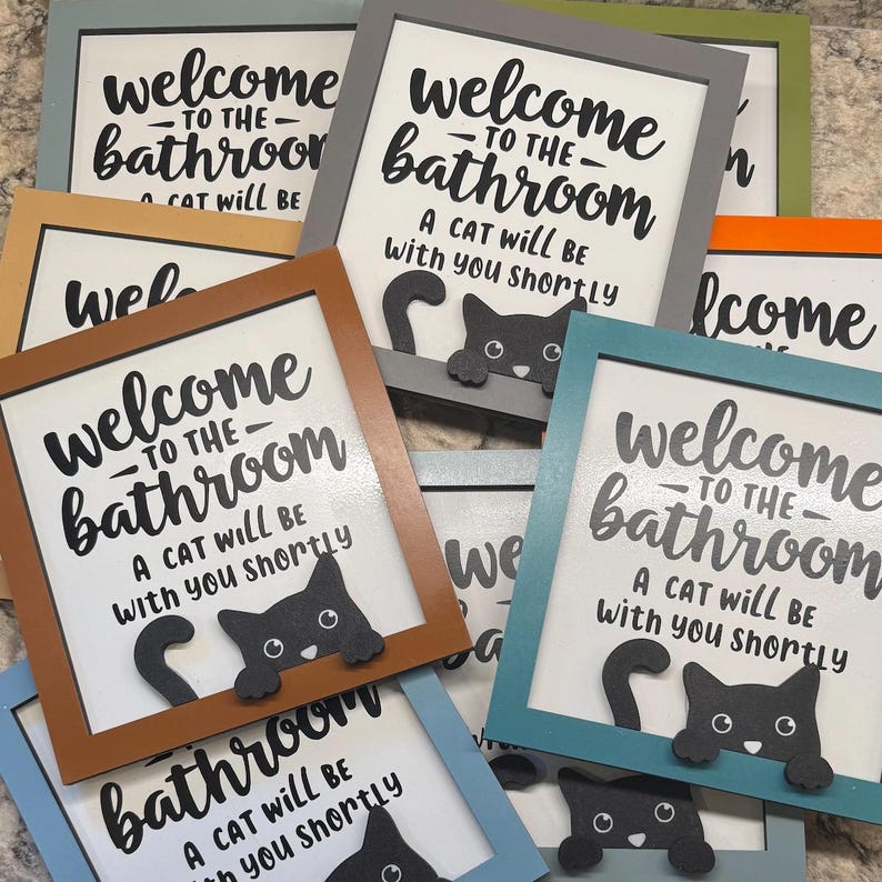 May include: A collection of framed bathroom signs with a playful cat design. Each sign features the text "Welcome to the Bathroom" and "A cat will be with you shortly" in black lettering. The frames come in various colors.
