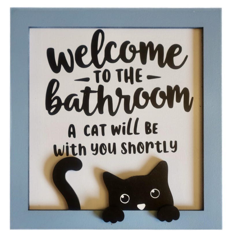 May include: A framed sign with a light blue border. The sign reads "Welcome to the Bathroom" and "A cat will be with you shortly." A black cat graphic is at the bottom of the sign.