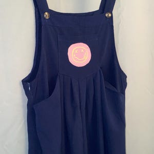 May include: A dark blue overall dress featuring a square neckline, wide shoulder straps with metallic buttons, and a central chest pocket. The pocket displays a circular pink patch with a yellow embroidered smiley face graphic with X eyes.