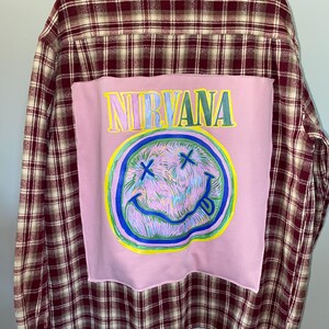 May include: A long-sleeve button-down shirt featuring a maroon and beige plaid pattern. A large, rectangular light pink fabric patch is sewn onto the back, displaying the word NIRVANA in yellow and a hand-drawn, multi-colored smiley face graphic.