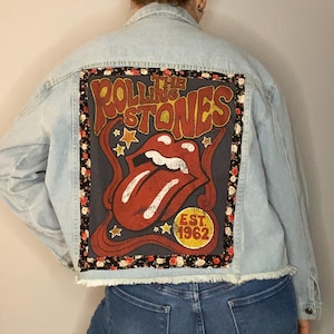 May include: Light blue denim jacket with a large, rectangular patch on the back featuring The Rolling Stones logo. The patch has a black border with a floral pattern and the band's iconic tongue and lips design in red.
