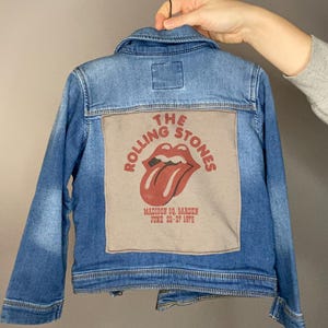 May include: A light blue denim jacket with a graphic on the back featuring the Rolling Stones logo and text. The graphic includes the band's iconic tongue and lips design, along with "Madison Sq. Garden June 22-27 1975."